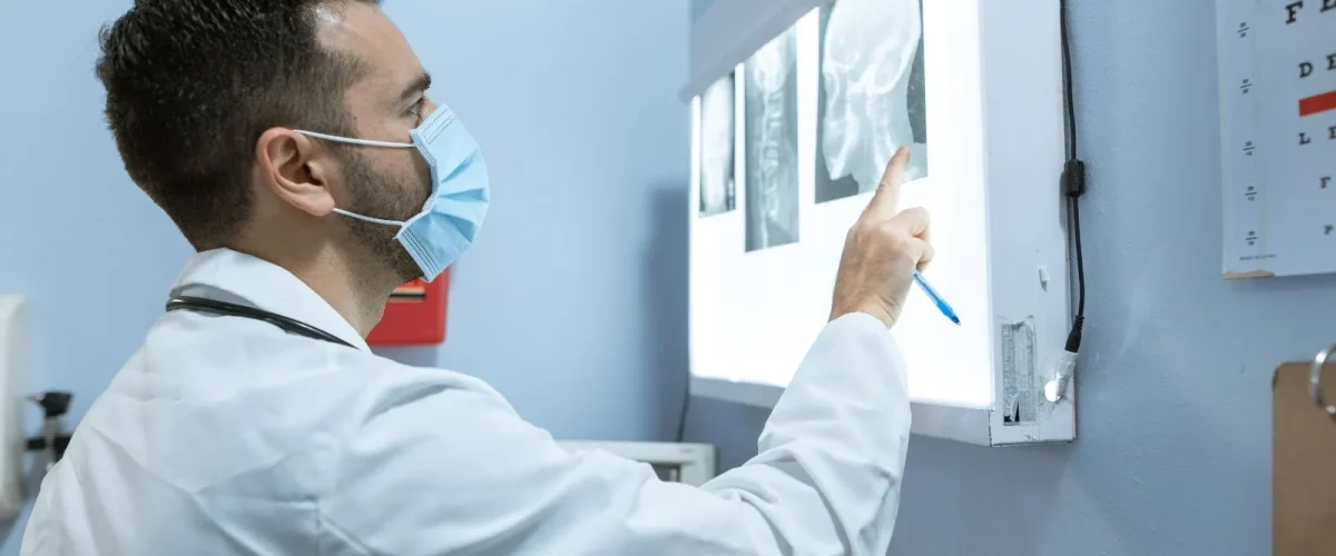 Doctor in a mask reviewing X-ray images on a light board, assessing diagnostic scans.